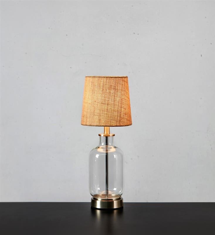 Lampa stołowa z przezroczystą szklaną podstawą i abażurem w kolorze beżowym, ustawiona na ciemnym blacie, na tle jasnej ściany.