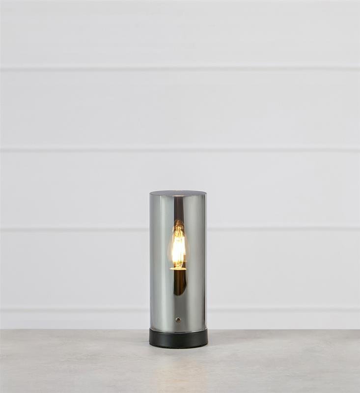 Lampa stołowa o cylindrycznym kształcie z metalicznym wykończeniem, stojąca na jasnym tle.