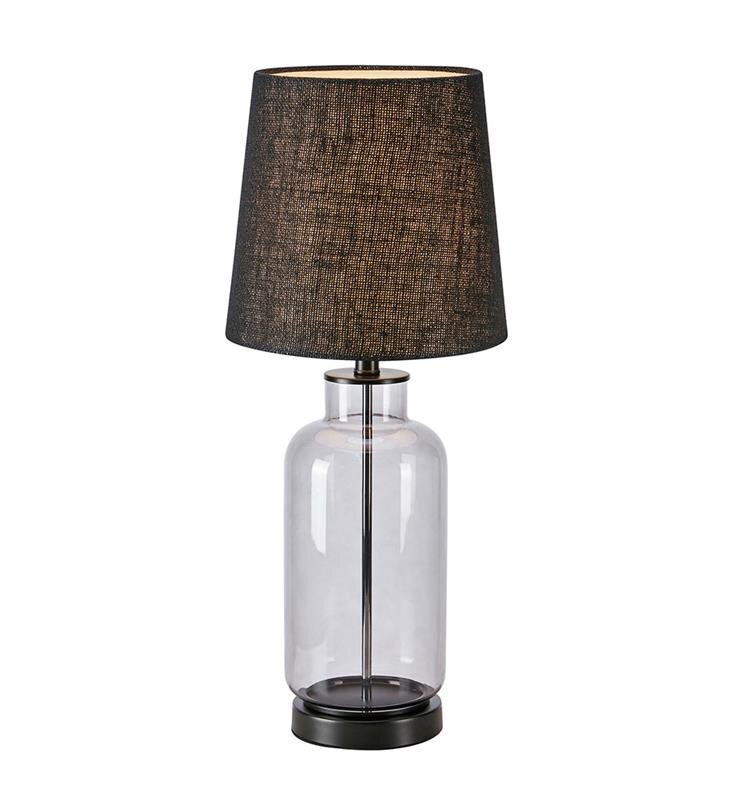 Lampa stołowa z przezroczystą szklaną podstawą i ciemnym abażurem o teksturowanej powierzchni. Podstawa lampy jest cylindryczna, a abażur ma kształt stożka.