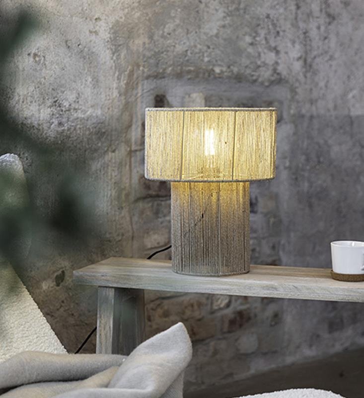 Lampa stołowa Markslöjd SOGA 108769 o cylindrycznym kształcie z jasnym abażurem stoi na drewnianym stoliku przy ścianie z surowej cegły.