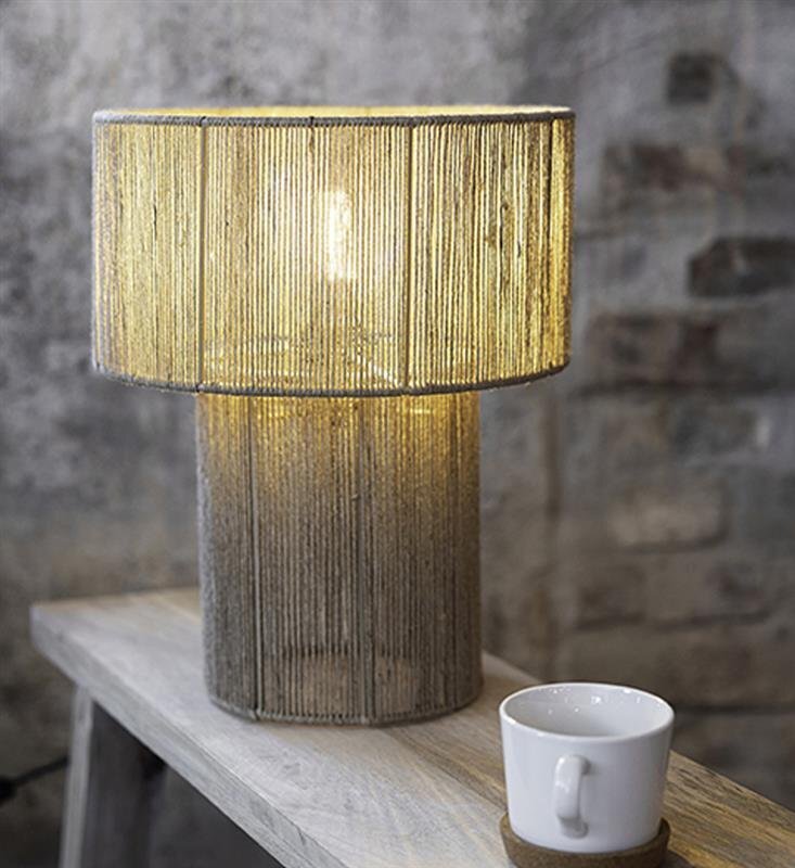 Stylowa lampa stołowa z abażurem z jasnego materiału stoi na drewnianym stole obok białej filiżanki na podstawce.