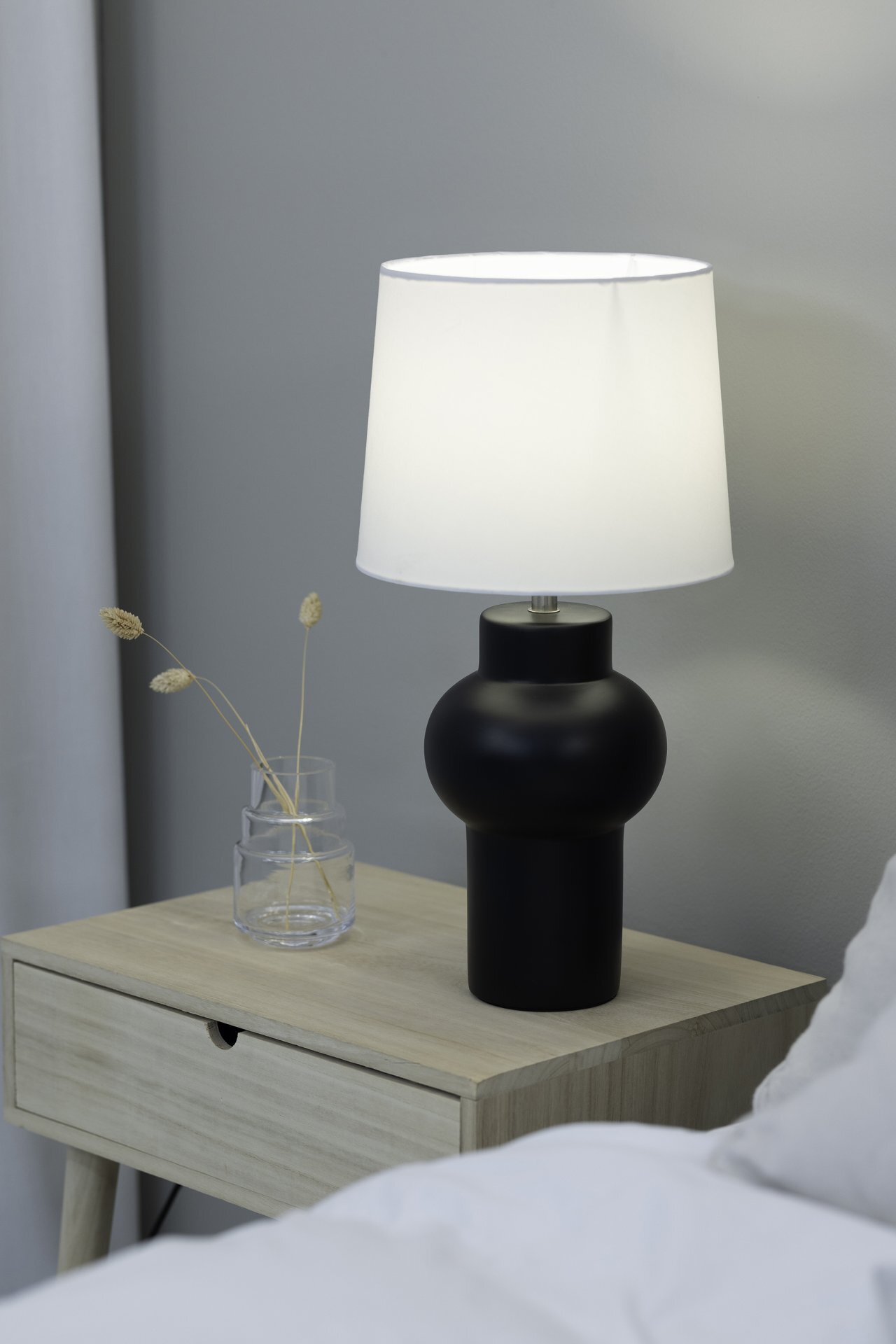 Na drewnianym stoliku nocnym stoi czarna lampa stołowa z białym abażurem. Obok lampy znajduje się szklany wazon z kilkoma suchymi gałązkami.