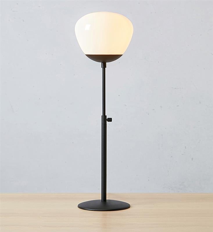 Lampa stołowa z białym kloszem i czarną podstawą, ustawiona na drewnianej powierzchni, na tle szarej ściany.