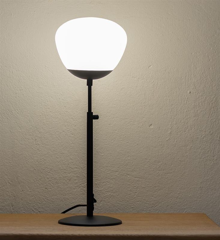 Lampa stołowa z białym kloszem i czarną podstawą stoi na drewnianej powierzchni, oświetlając ścianę w tle.
