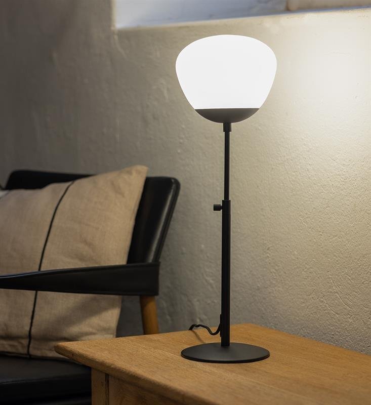 Lampa stołowa z białym kloszem i czarną podstawą stoi na drewnianym stoliku obok fotela z poduszką.