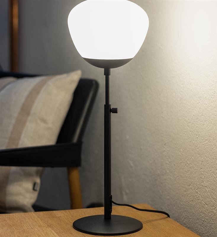 Lampa stołowa z białym kloszem i czarną podstawą stoi na drewnianym stole, w tle widoczna jest część fotela z poduszką.