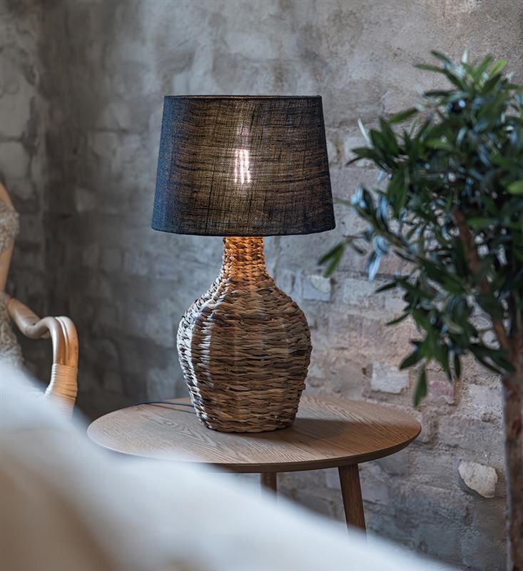 Na drewnianym stoliku stoi lampa stołowa z plecioną podstawą i ciemnym abażurem. W tle widoczna jest ceglana ściana oraz fragment rośliny.