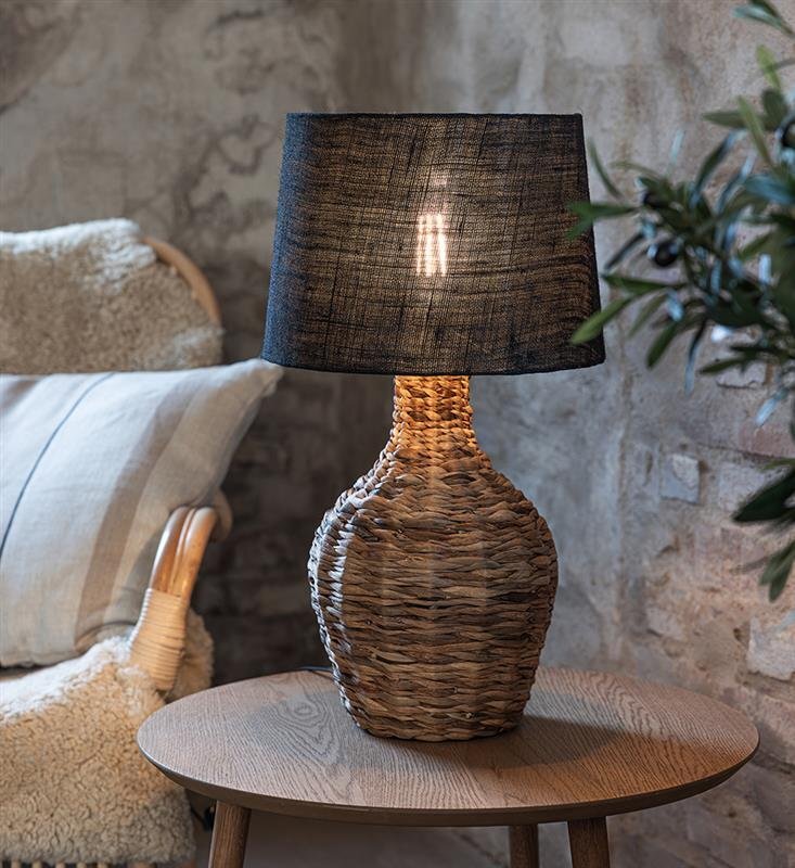 Na drewnianym stoliku stoi lampa stołowa z plecioną podstawą i ciemnym abażurem, przez który prześwituje światło. W tle widoczna jest ściana z cegieł oraz fragment fotela z poduszką.