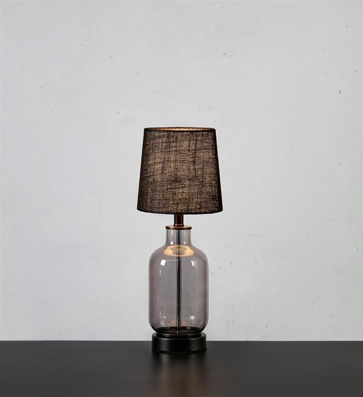 Lampa stołowa z ciemnym abażurem i przezroczystą podstawą stoi na ciemnym blacie, na tle jasnej ściany.