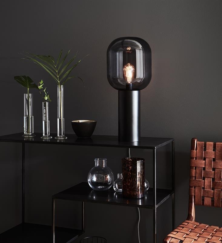 Czarna lampa stołowa o nowoczesnym designie stoi na ciemnym stoliku obok wazonów z roślinami i szklanych naczyń, na tle ciemnej ściany.