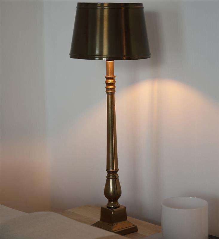 Lampa stołowa z metalowym kloszem i ozdobną podstawą stoi na drewnianym stoliku, obok znajduje się biały kubek.