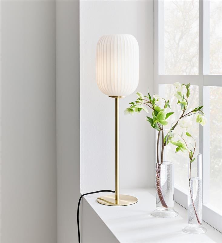 Na zdjęciu znajduje się elegancka lampa stołowa z białym, żebrowanym kloszem i złotą podstawą, ustawiona na parapecie obok dwóch wazonów z zielonymi gałązkami. W tle widoczne jest okno z delikatnie rozmytym widokiem na zewnątrz.