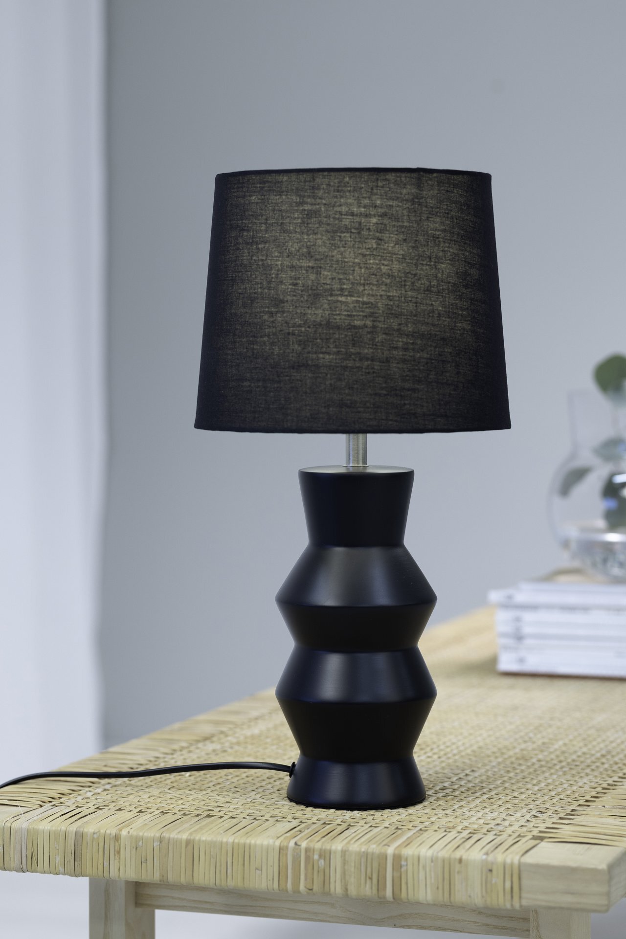 Czarna lampa stołowa z geometryczną podstawą i prostym abażurem stoi na jasnym, plecionym stole. W tle widoczne są książki i szklany wazon.