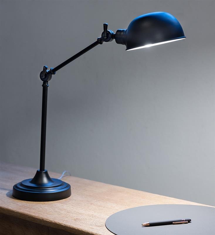 Czarna lampa biurkowa na wysięgniku stoi na drewnianym blacie, obok znajduje się okrągła podkładka z długopisem.