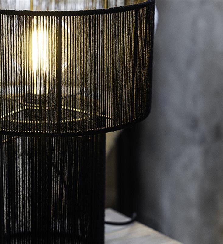 Lampa stołowa z czarnym, ażurowym kloszem, przez który prześwituje światło żarówki. Tło jest szare i niewyraźne.