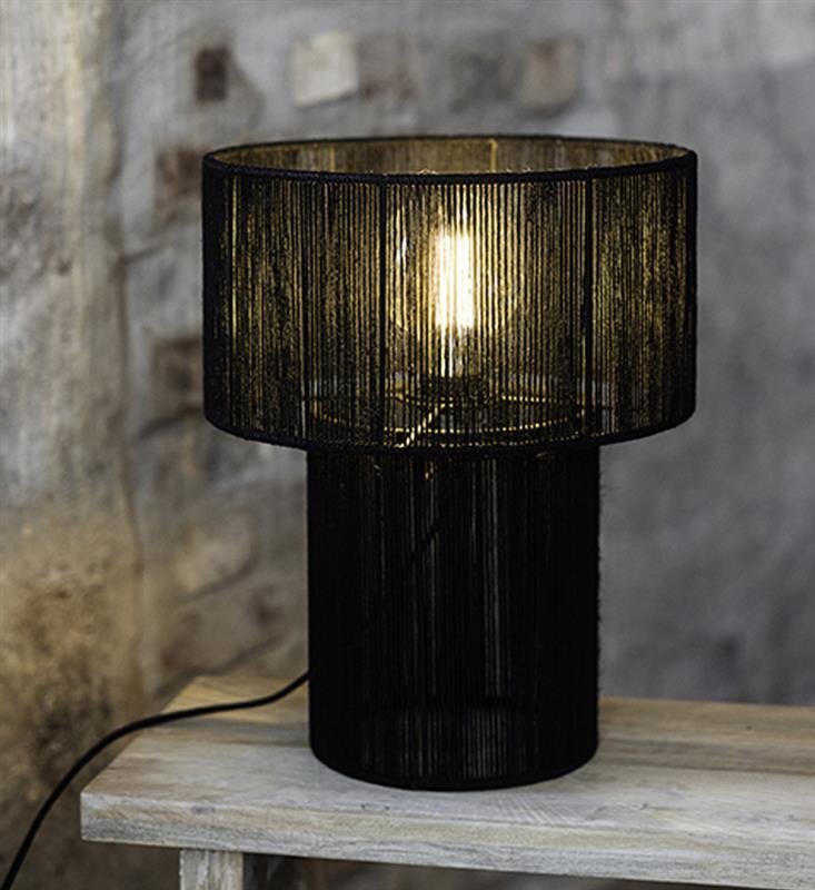 Czarna lampa stołowa z ażurowym kloszem, emitująca ciepłe światło, stoi na drewnianym blacie. W tle widoczna jest ceglana ściana.