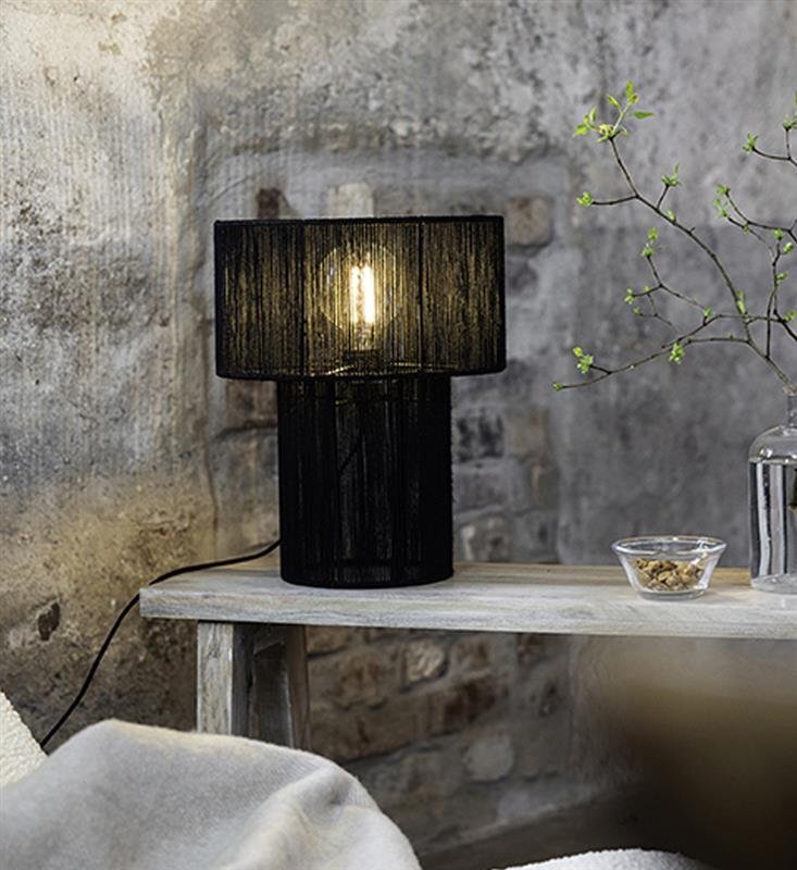 Czarna lampa stołowa z widoczną żarówką stoi na drewnianym stole przy ścianie z cegieł. Obok lampy znajduje się szklana miseczka z orzechami i gałązka w wazonie.