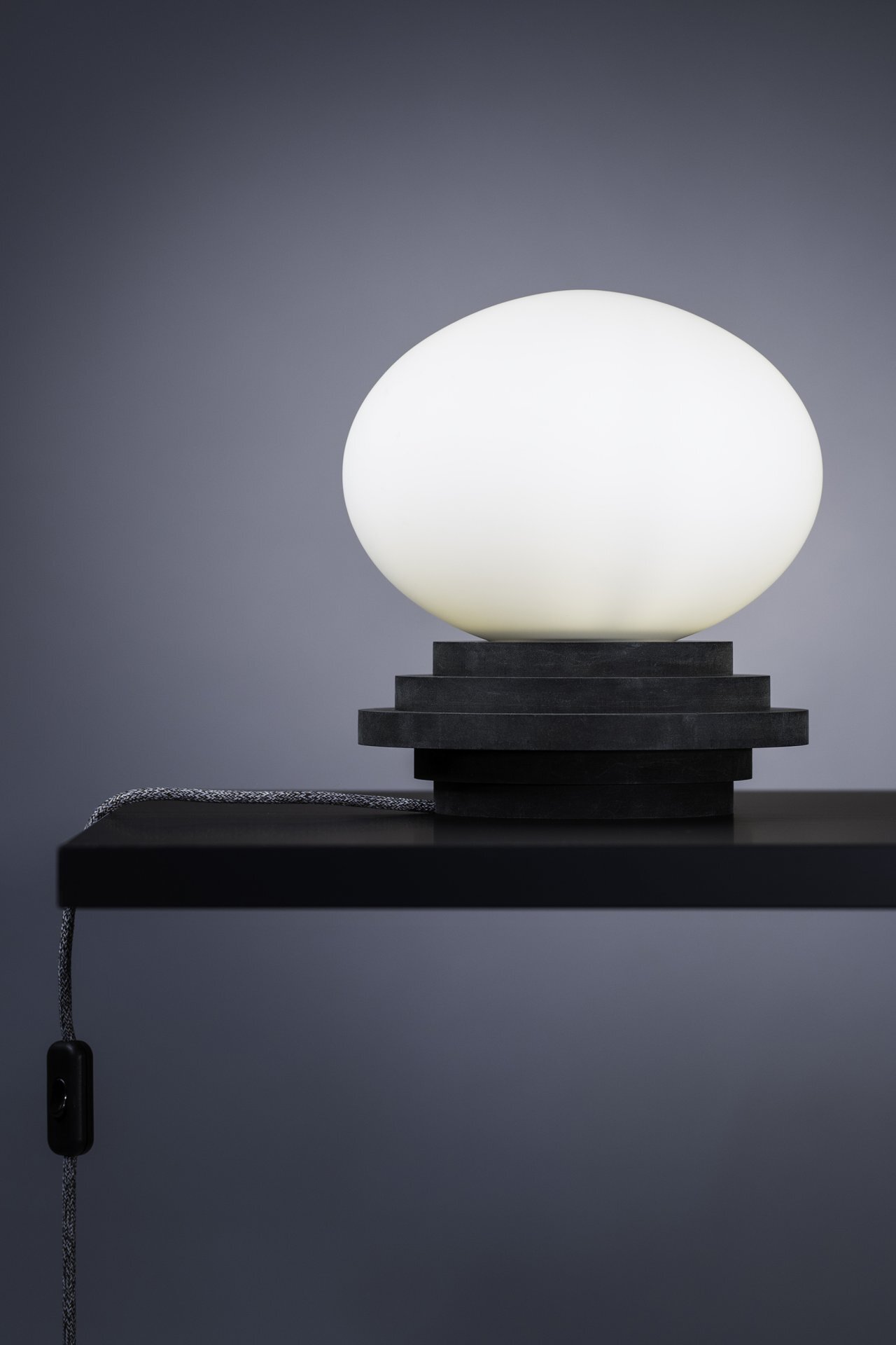 Na zdjęciu widoczna jest lampa stołowa z białym, owalnym kloszem umieszczonym na czarnym, geometrycznym postumencie. Lampa stoi na ciemnym blacie, a z boku zwisa przewód z włącznikiem.