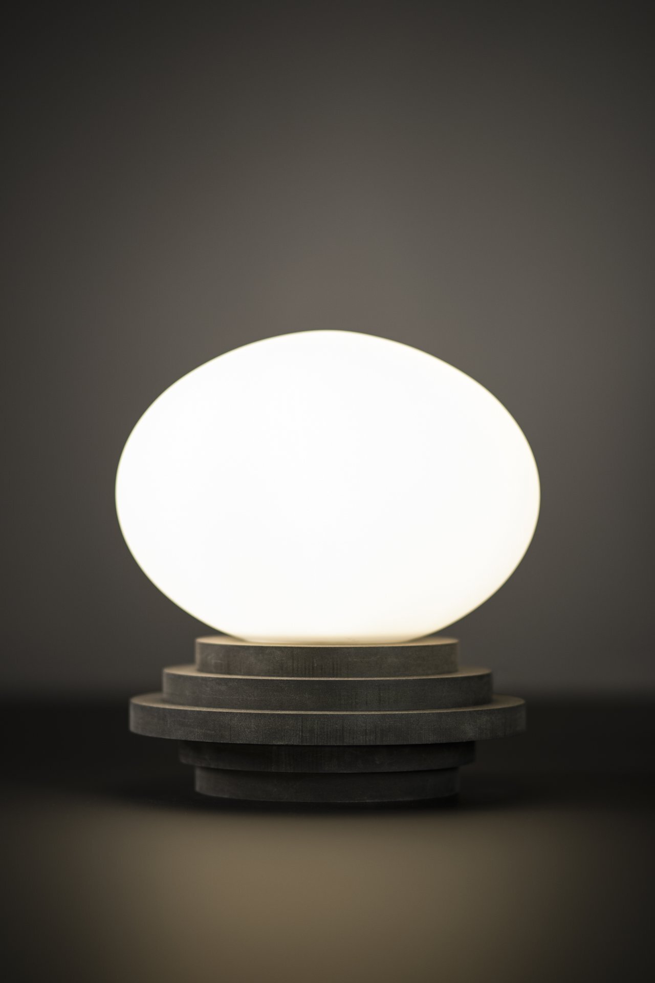 Lampa stołowa o owalnym, jasnym kloszu umieszczona na ciemnej, wielowarstwowej podstawie, na neutralnym tle.