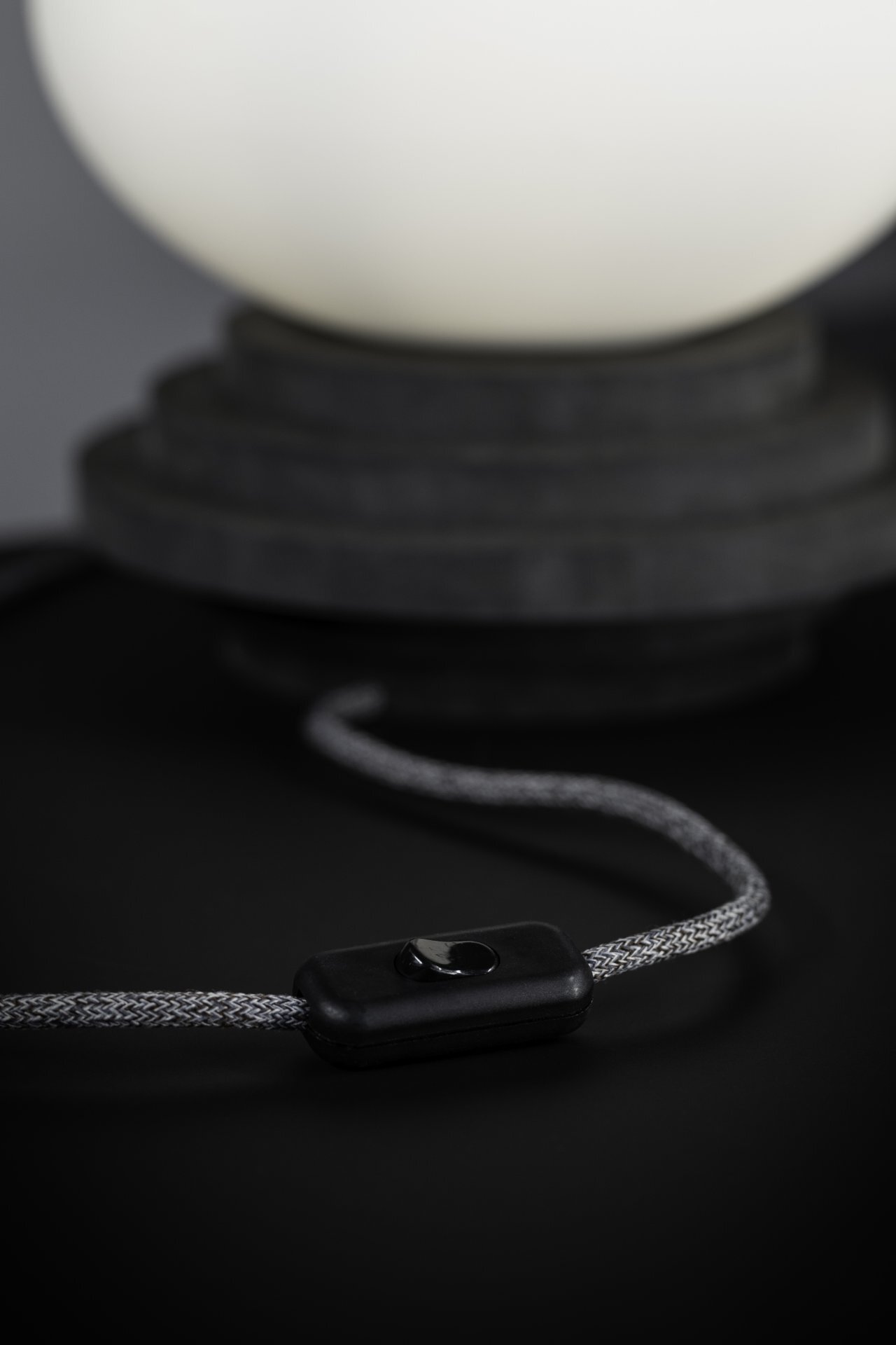 Zdjęcie przedstawia podstawę lampy stołowej z czarnym włącznikiem na szarym kablu, umieszczonym na ciemnym tle.