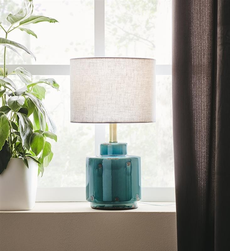 Lampa stołowa z niebieską ceramiczną podstawą i białym abażurem stoi na parapecie obok zielonej rośliny w białej doniczce, w tle widoczne jest okno z zasłoną.