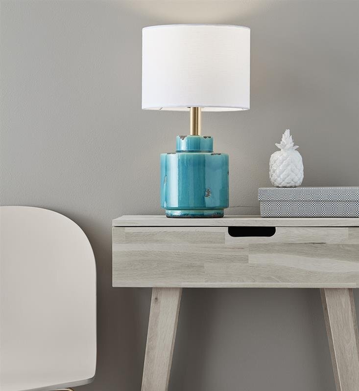 Na drewnianym stoliku stoi lampa stołowa z niebieską ceramiczną podstawą i białym abażurem. Obok lampy znajduje się dekoracyjny biały ananas i książka w szarym etui.