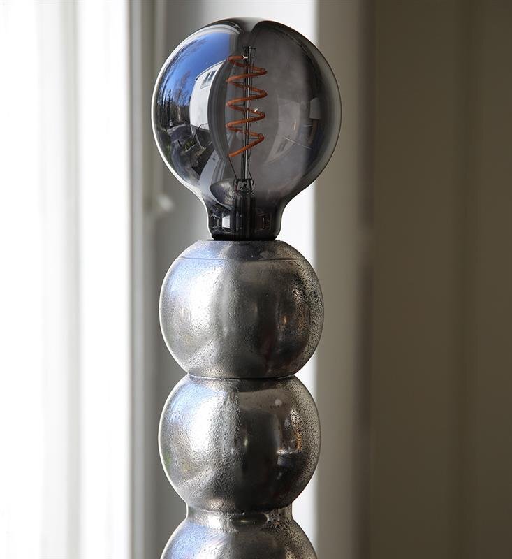 Lampa stołowa z metalową podstawą składającą się z kilku kulistych segmentów i dużą żarówką o widocznym żarniku. Tło jest jasne, z widocznym oknem po lewej stronie.