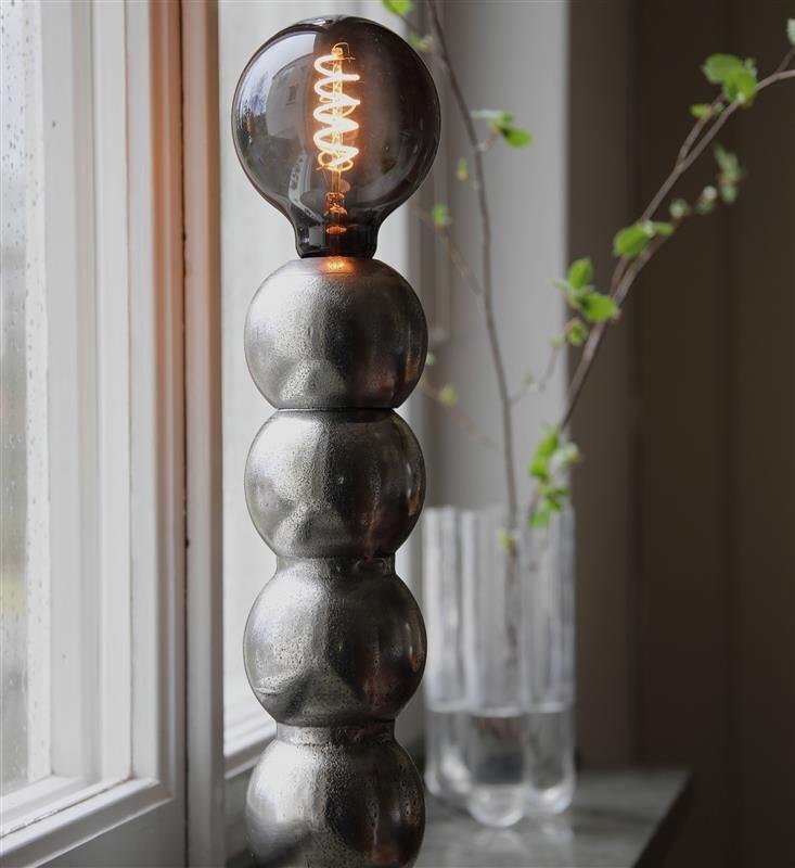 Lampa stołowa z metalową podstawą składającą się z kilku kulistych segmentów, zakończona żarówką z widocznym żarnikiem. W tle widać okno i przezroczysty wazon z gałązkami.