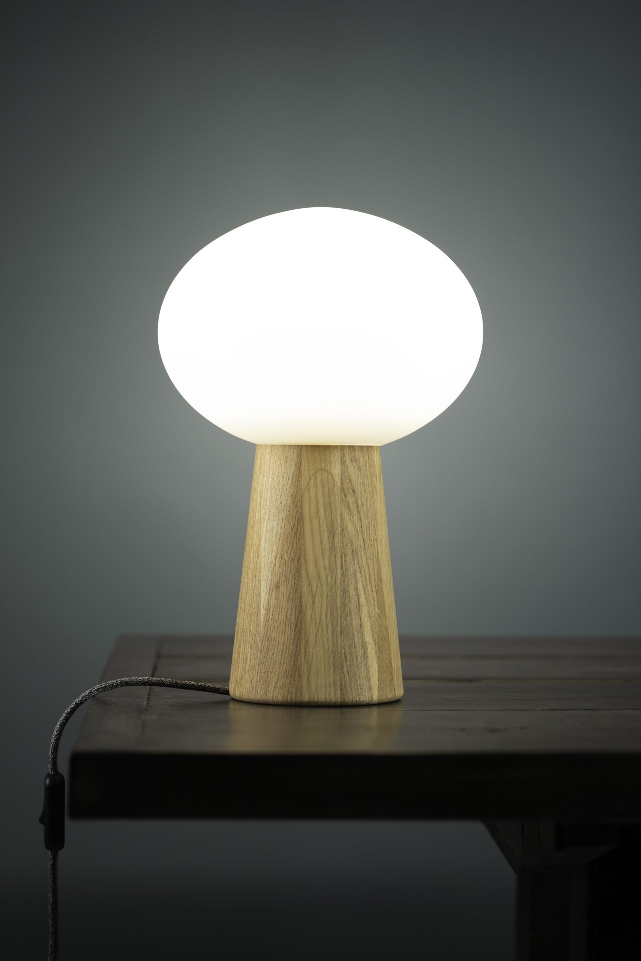 Lampa stołowa z drewnianą podstawą i białym, kulistym kloszem stoi na ciemnym stole, z widocznym przewodem zasilającym.