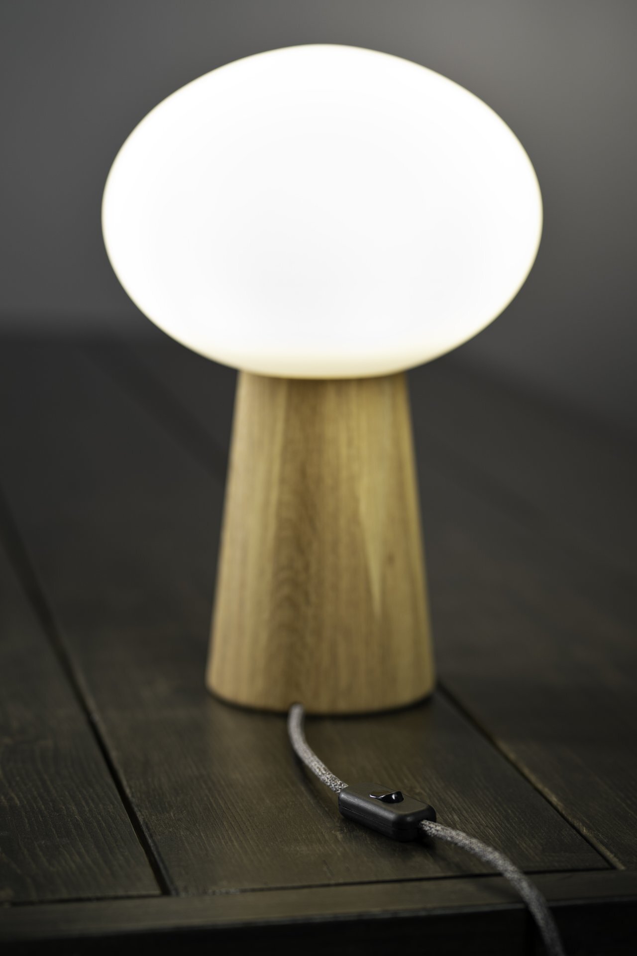 Lampa stołowa z drewnianą podstawą i białym, owalnym kloszem, stojąca na ciemnym drewnianym stole z widocznym przewodem z włącznikiem.