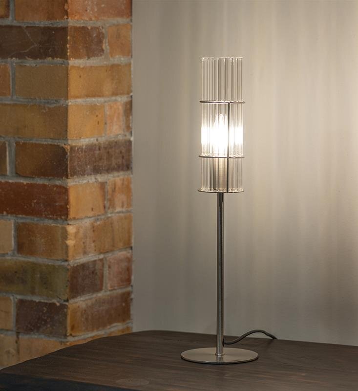 Lampa stołowa z metalową podstawą i szklanym kloszem stoi na ciemnym drewnianym stole, obok ściany z cegieł.