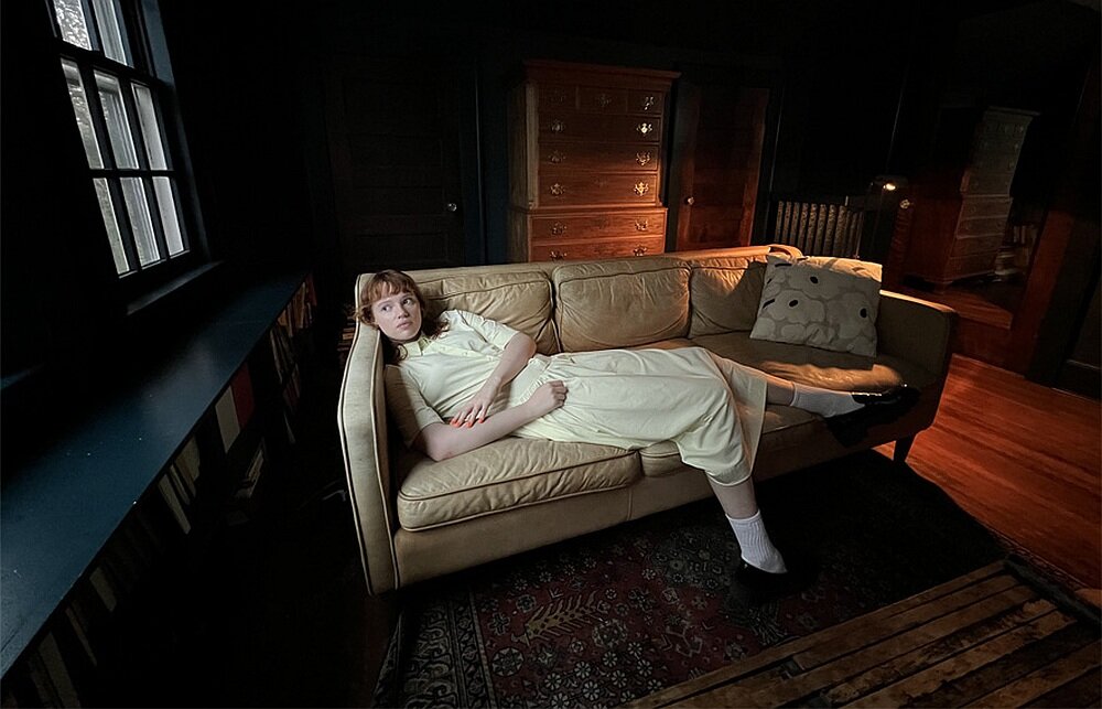 Osoba leży na jasnej kanapie w ciemnym pokoju, obok poduszka. W tle widoczne są drewniane meble i dywan na podłodze.