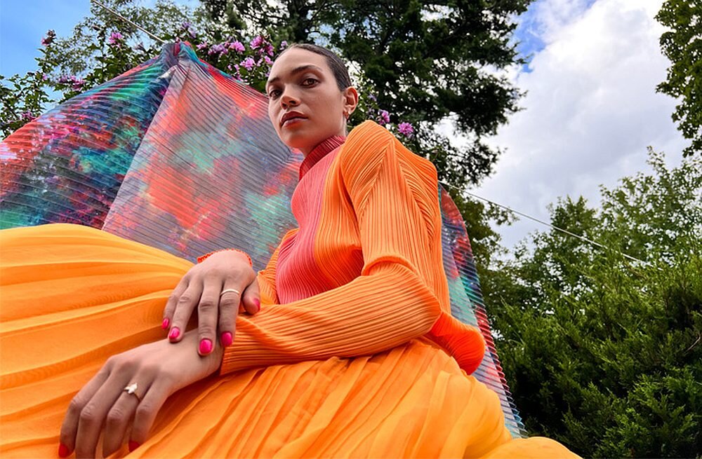 Osoba w pomarańczowym stroju siedzi na tle kolorowej tkaniny i zieleni. W tle widać drzewa i niebo z chmurami.