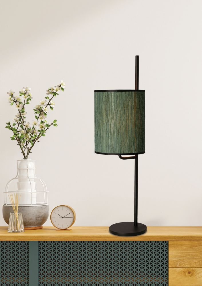 Lampa stołowa z zielonym abażurem stoi na drewnianym blacie obok wazonu z kwiatami i zegara. Tło jest jednolite i jasne.
