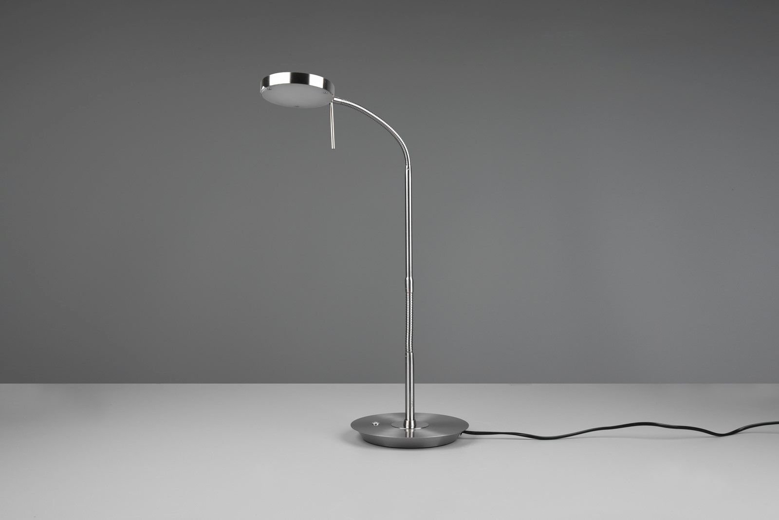 Na zdjęciu znajduje się nowoczesna lampa biurkowa LED o metalicznym wykończeniu, stojąca na jasnym tle z widocznym przewodem zasilającym.