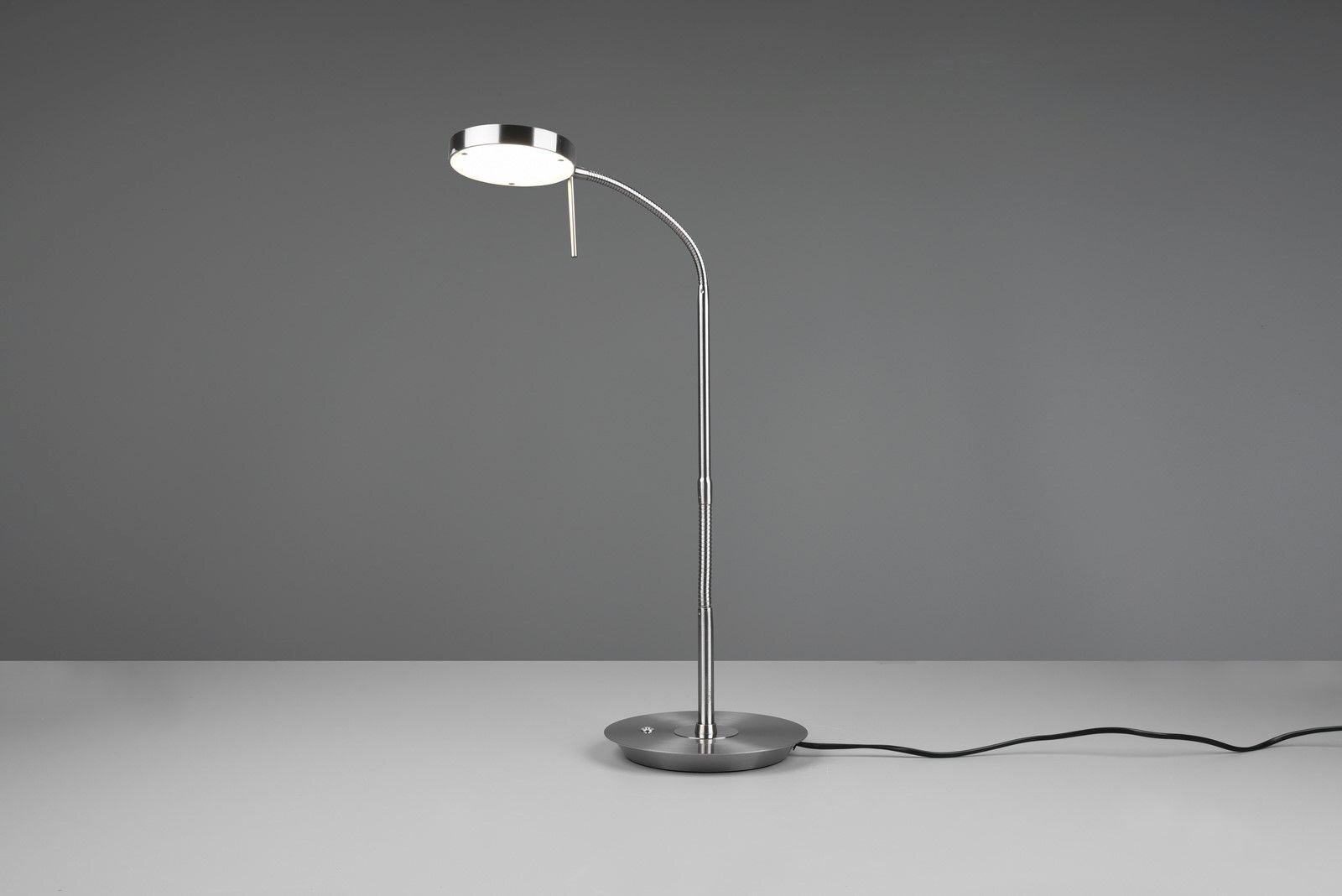 Na zdjęciu znajduje się nowoczesna lampa biurkowa LED o metalicznym wykończeniu, stojąca na jasnym tle z widocznym przewodem zasilającym.