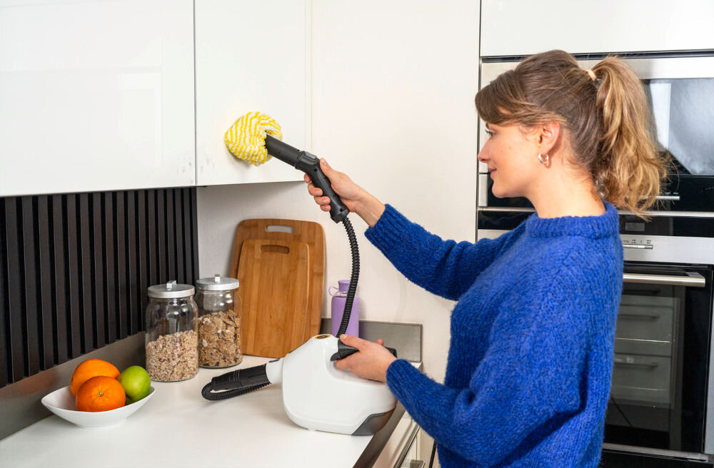 Osoba używa czyścika parowego do czyszczenia białej szafki kuchennej. Na blacie znajdują się owoce w misce, słoiki z produktami spożywczymi i deska do krojenia.