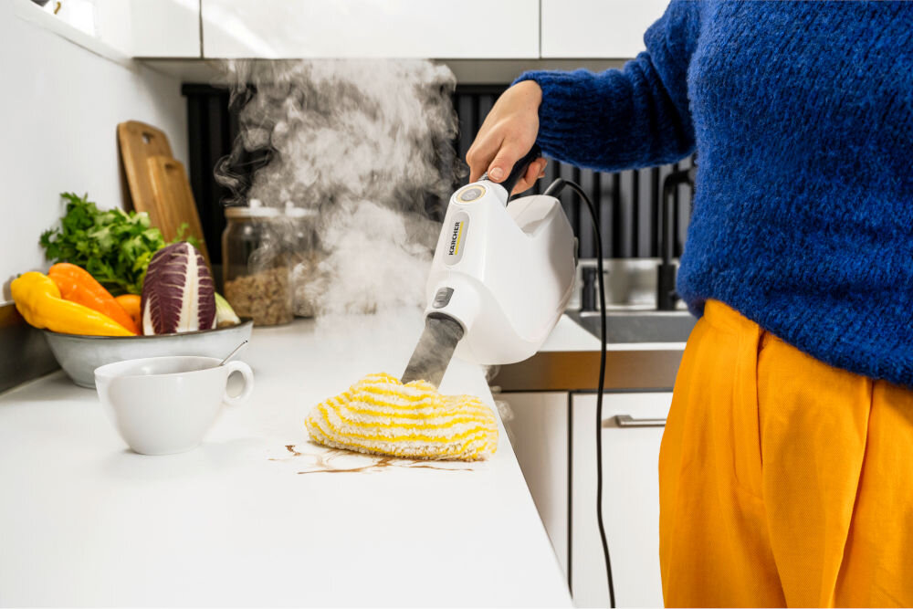 Osoba używa białego czyścika parowego z logo \'KARCHER\' do czyszczenia blatu kuchennego. Na blacie znajduje się żółto-biała ściereczka, a w tle widoczne są warzywa i filiżanka.