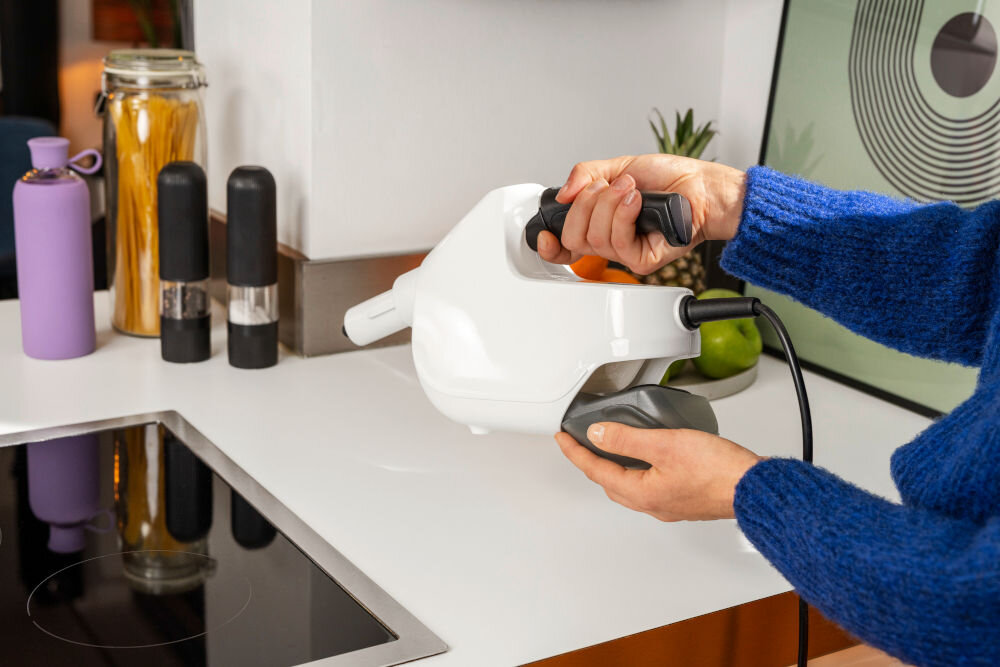 Osoba trzyma biały czyścik parowy na blacie kuchennym. W tle widoczne są różne przedmioty kuchenne, takie jak przyprawniki i butelka.