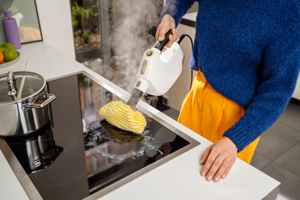 Osoba używa białego czyścika parowego do czyszczenia płyty kuchennej. Na płycie znajduje się żółto-biała nakładka czyszcząca, a obok stoi garnek ze szklaną pokrywką.