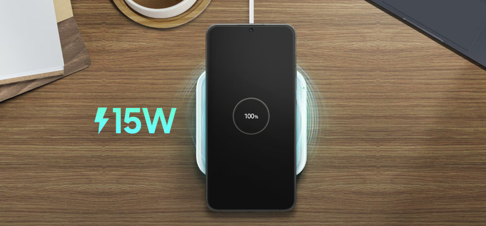 Na drewnianym blacie znajduje się smartfon leżący na białej ładowarce indukcyjnej, z wyświetlaczem pokazującym 100% naładowania. Obok widoczny jest symbol błyskawicy i napis \'15W\'.
