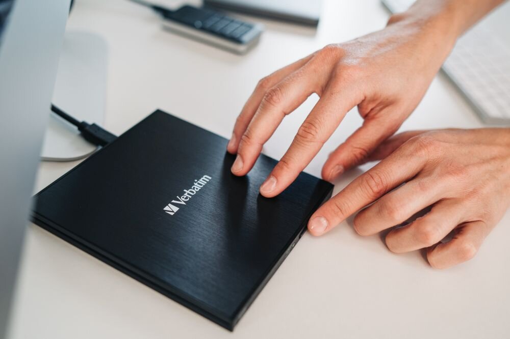 Napęd VERBATIM 98938 Zbliżenie na dłonie zamykające czarny napęd optyczny podłączony do komputera stojącego na biurku zewnętrzna nagrywarka USB