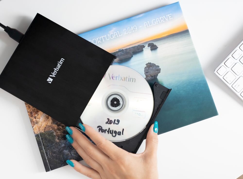 Napęd VERBATIM 98938 Czarny napęd optyczny z wysuniętą tacką, w której znajduje się płyta z napisem '2019 Portugal', pod spodem leży album ze zdjęciem krajobrazu nadmorskiego, obok fragment klawiatury zasilanie z USB, idealna do notebooków