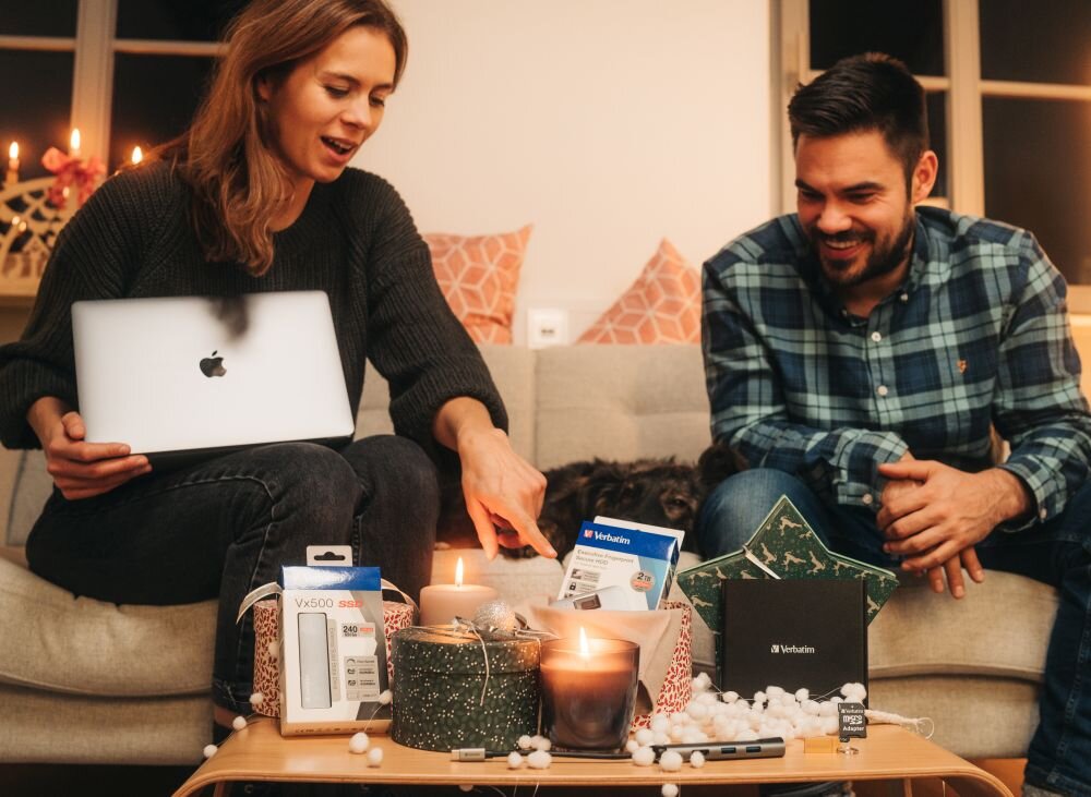 Napęd VERBATIM 98938 Kobieta z laptopem i mężczyzna w koszuli w kratę siedzą przy stole, na którym stoją zapalone świece, opakowania z akcesoriami komputerowymi i czarny napęd optyczny archiwizacja danych na lata, kompatybilna z Windows i MacOS