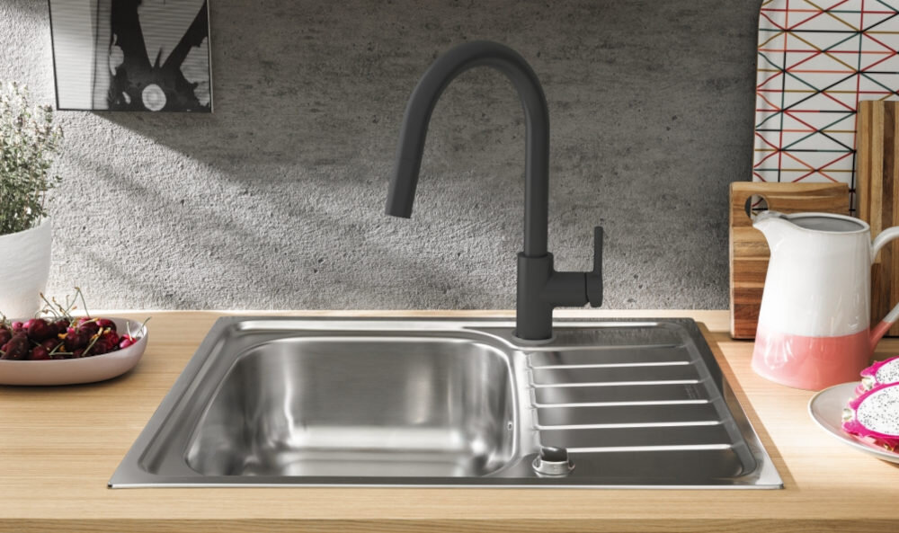 Na obrazie widoczny jest zlew kuchenny z metalową komorą i ociekaczem, wyposażony w czarną baterię kuchenną z wyciąganą wylewką. W tle znajdują się akcesoria kuchenne, takie jak deski do krojenia i dzbanek.
