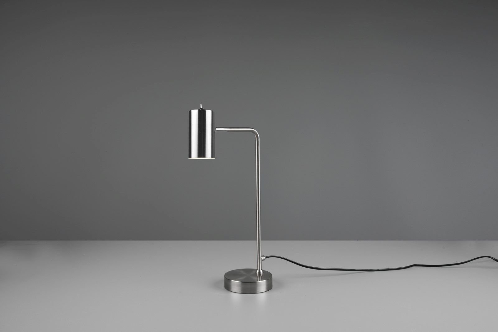 Srebrna lampa biurkowa o nowoczesnym designie stoi na szarej powierzchni z widocznym czarnym kablem zasilającym.