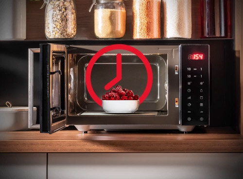 Otwarta kuchenka mikrofalowa z miseczką czerwonych owoców w środku, na drzwiach widoczny jest czerwony symbol zegara. Na panelu sterowania wyświetla się godzina 18:54.