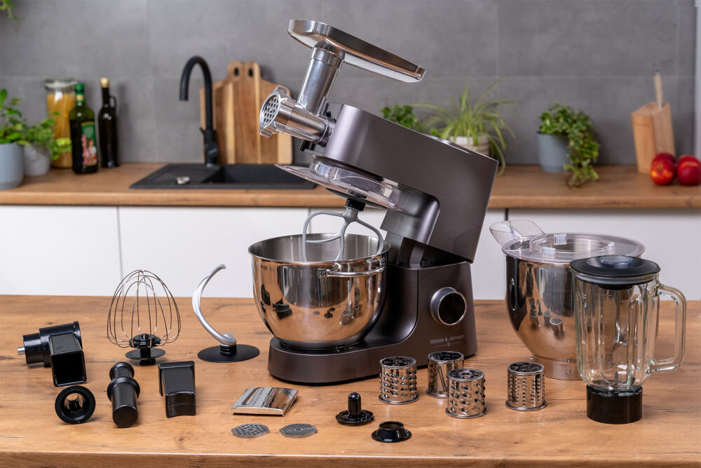 Na drewnianym blacie znajduje się robot kuchenny planetarny z metalową misą i różnymi akcesoriami, w tym trzepaczką, hakami do ciasta, tarkami i blenderem kielichowym. W tle widoczna jest kuchnia z zlewem, roślinami i przyborami kuchennymi.