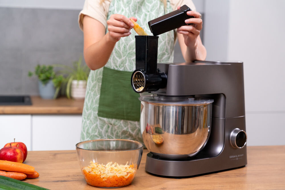 Osoba używa robota kuchennego planetarnego do szatkowania warzyw, które wpadają do miski. Na blacie znajdują się starte warzywa w szklanej misce oraz jabłka i marchewki.