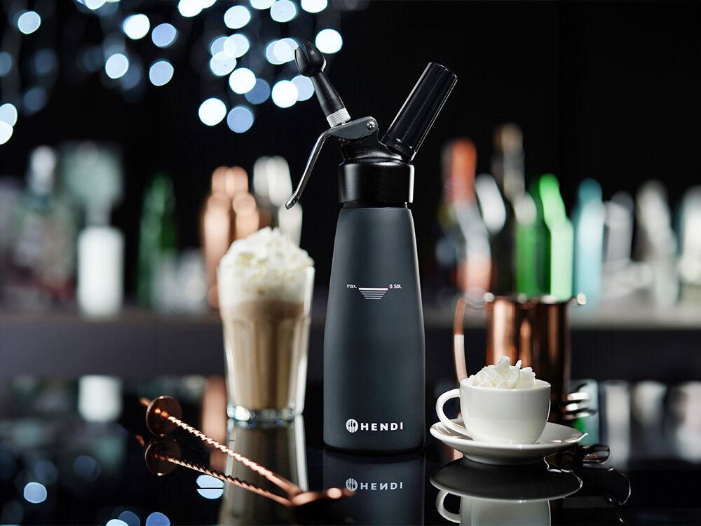 Na obrazie widoczny jest czarny syfon do bitej śmietany z logo \'HENDI\' oraz oznaczeniem \'MAX. 0.5L\'. W tle znajdują się rozmyte kształty butelek i filiżanka z bitą śmietaną na spodku.